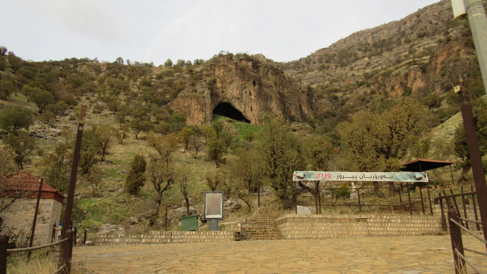 a wide dark cave mouth in a rocky hillside, with a paved terrace and a banner below it and scattered trees on the slope