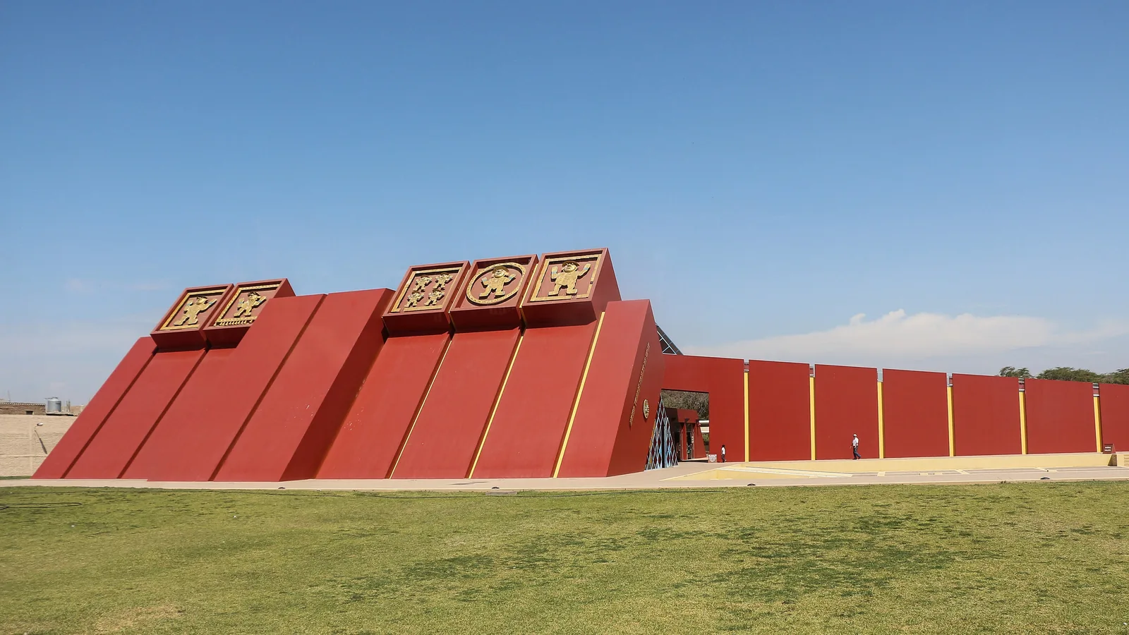 Angular red pyramid-shaped museum building with gold relief panels, seen from a grass lawn