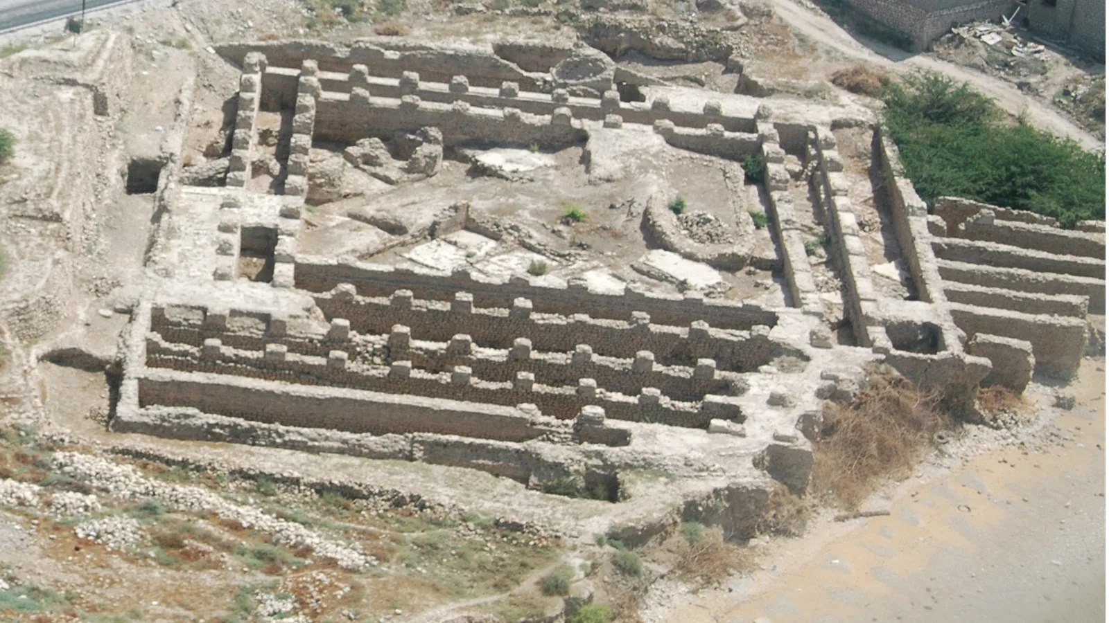 Aerial view of ruined mud-brick mosque walls with buttressed piers on a coastal hillside