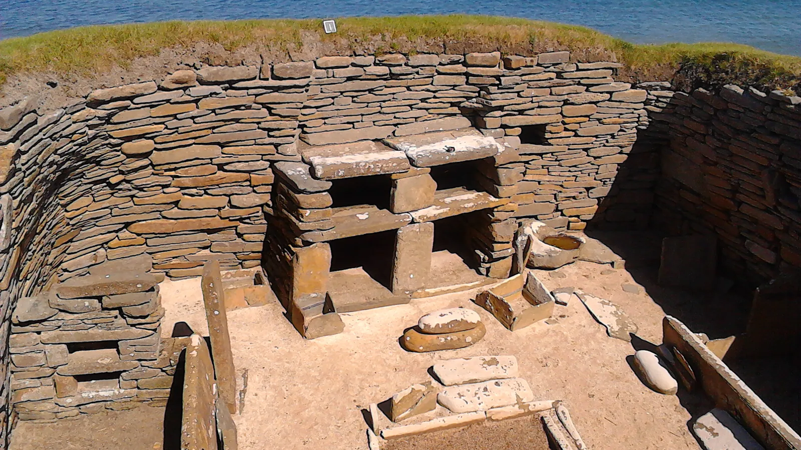 Interior of a drystone Neolithic house seen from above, with a two-shelf stone dresser against the far wall