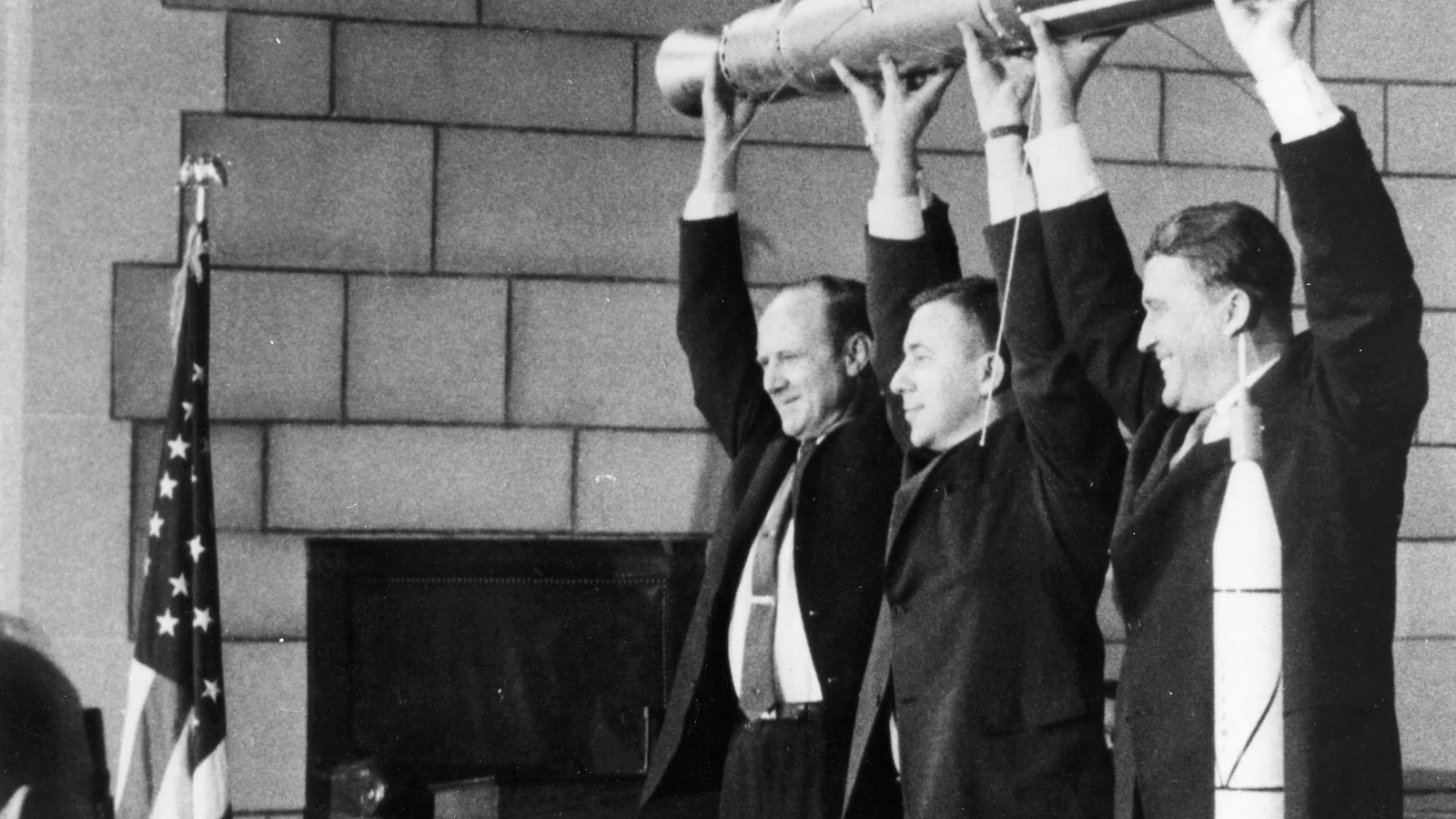 three men in suits holding up a small torpedo-shaped satellite model above their heads before reporters
