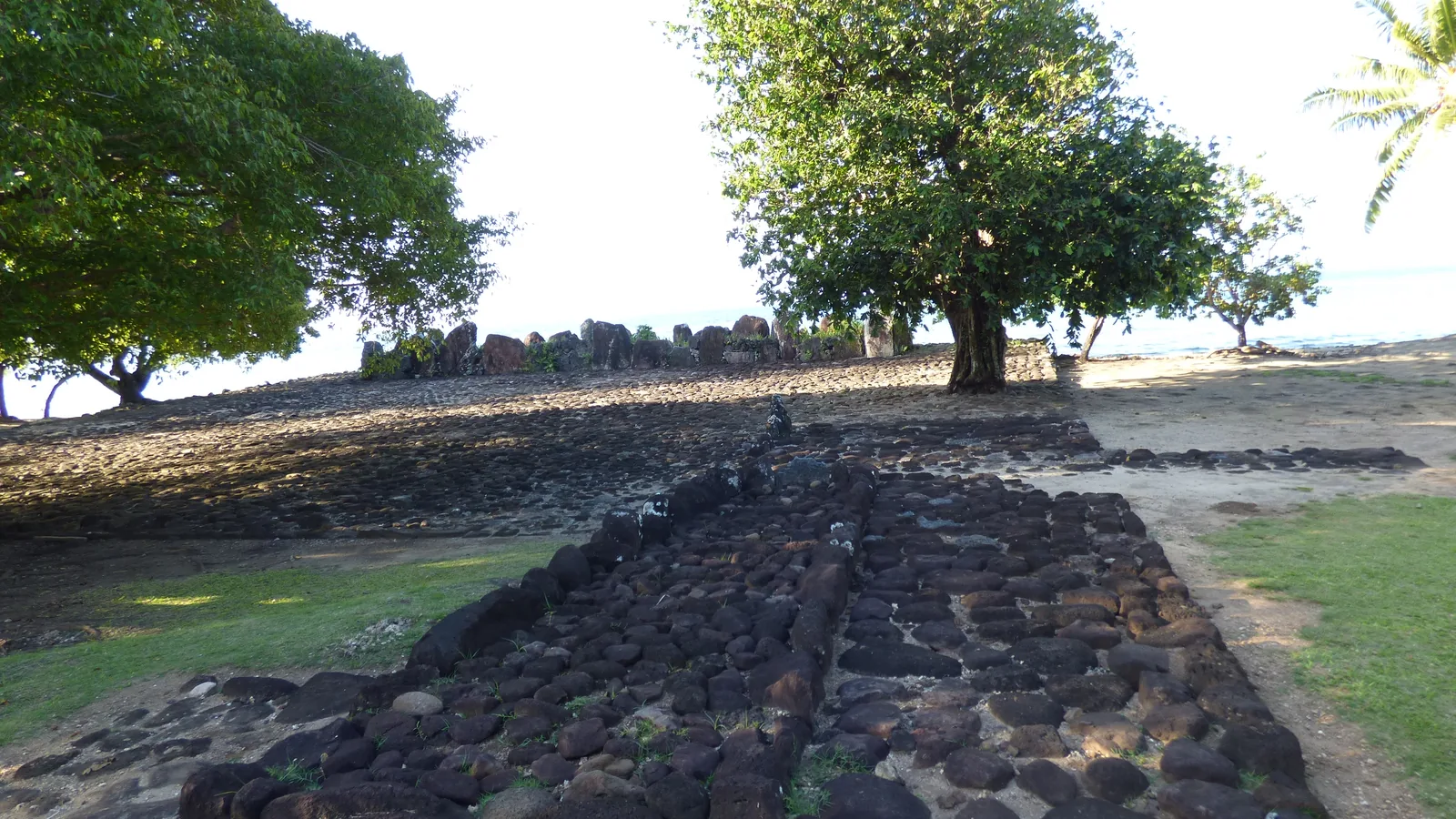 Close view of a coral-stone marae platform with low walls, a shade tree, and calm lagoon water beyond.