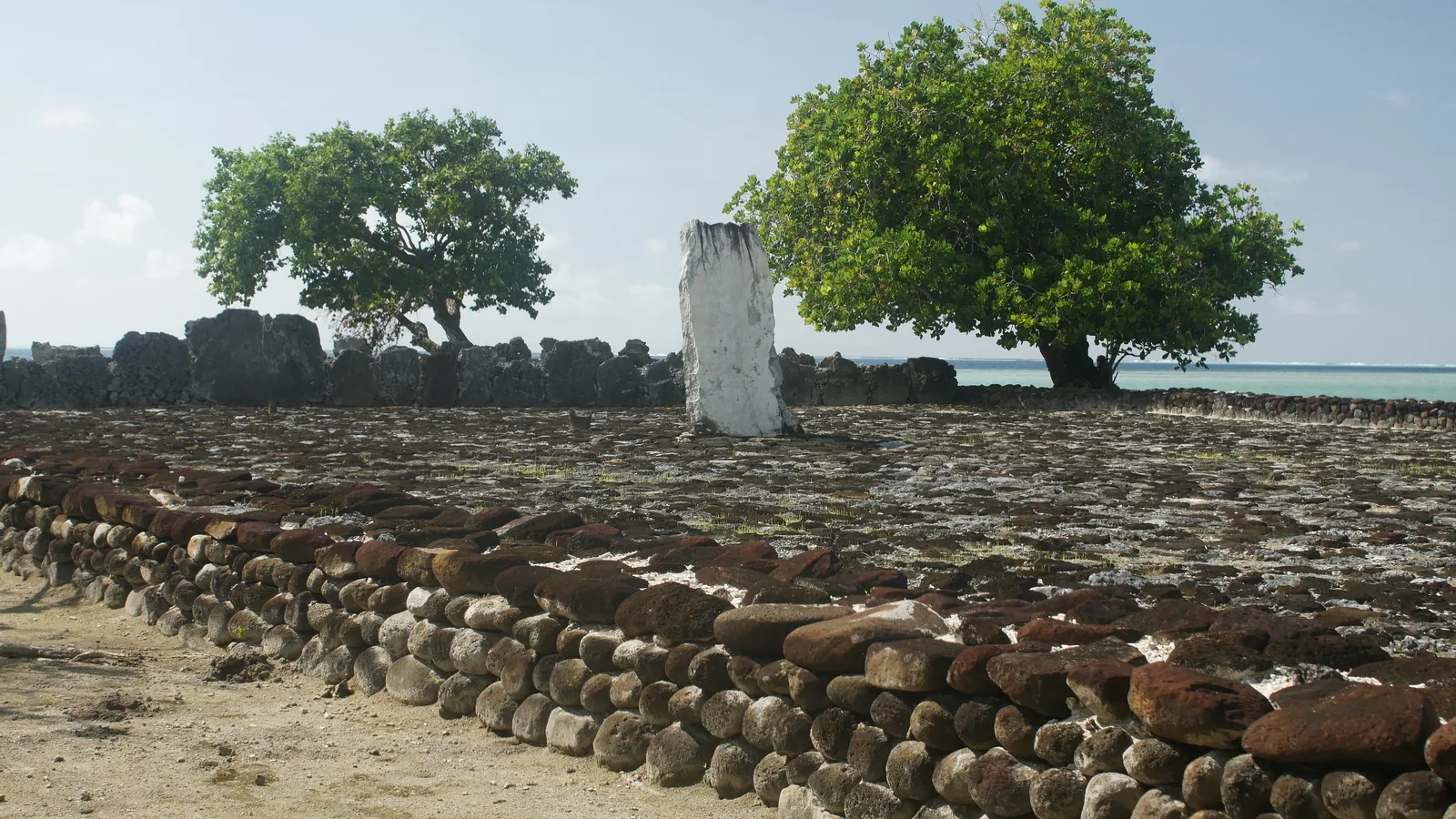 Stone-paved marae platform by a lagoon, with a low coral wall, an upright stone slab, and shade trees.
