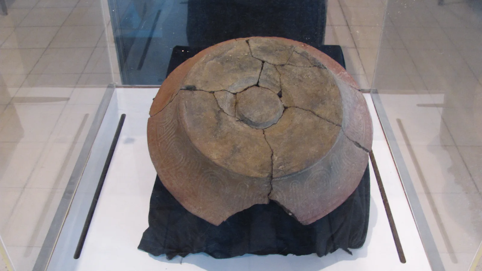 a reconstructed brown clay pot with a wide shoulder and dentate-stamped bands, displayed in a glass case
