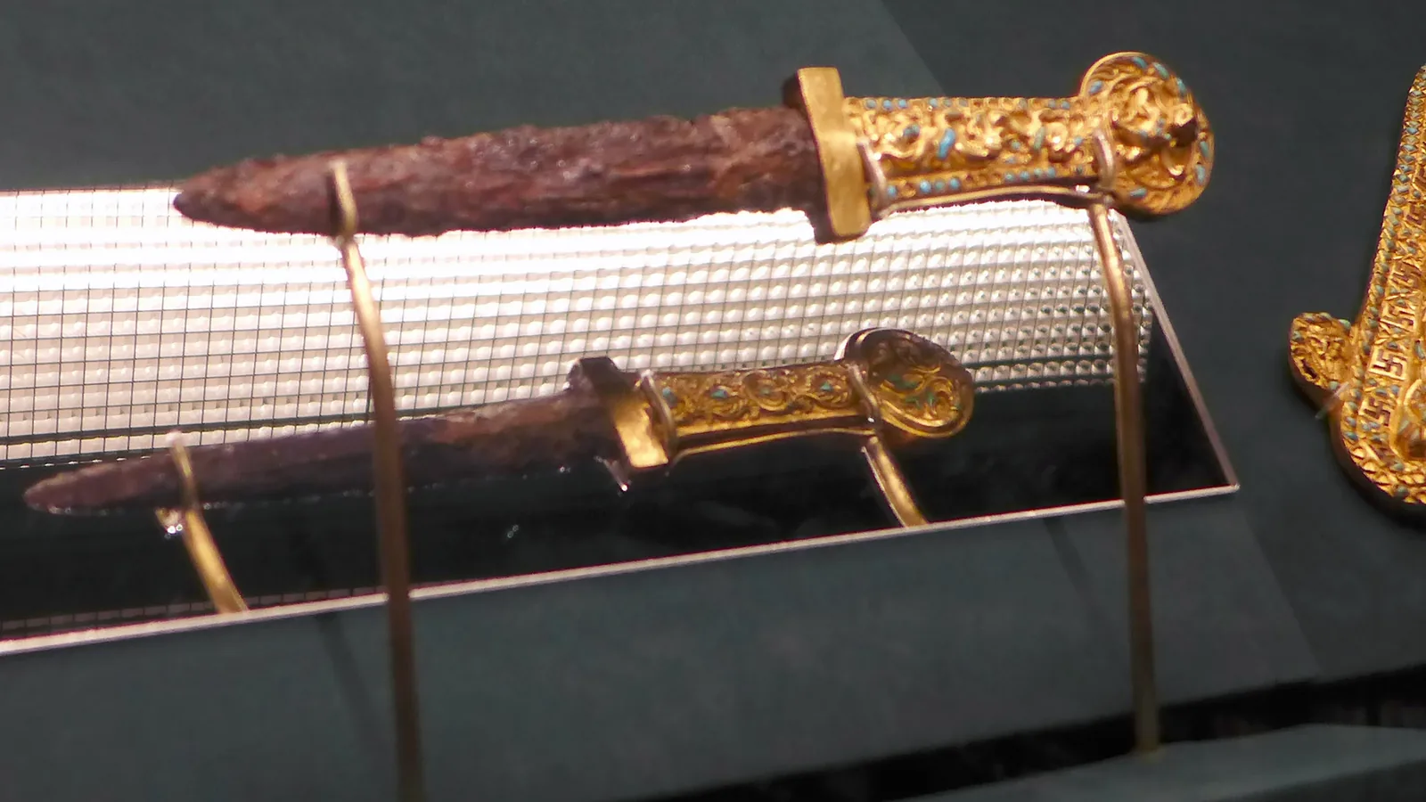 two iron daggers with gold hilts set with turquoise, and a gold scabbard ornament decorated with coiled creatures, in a museum case