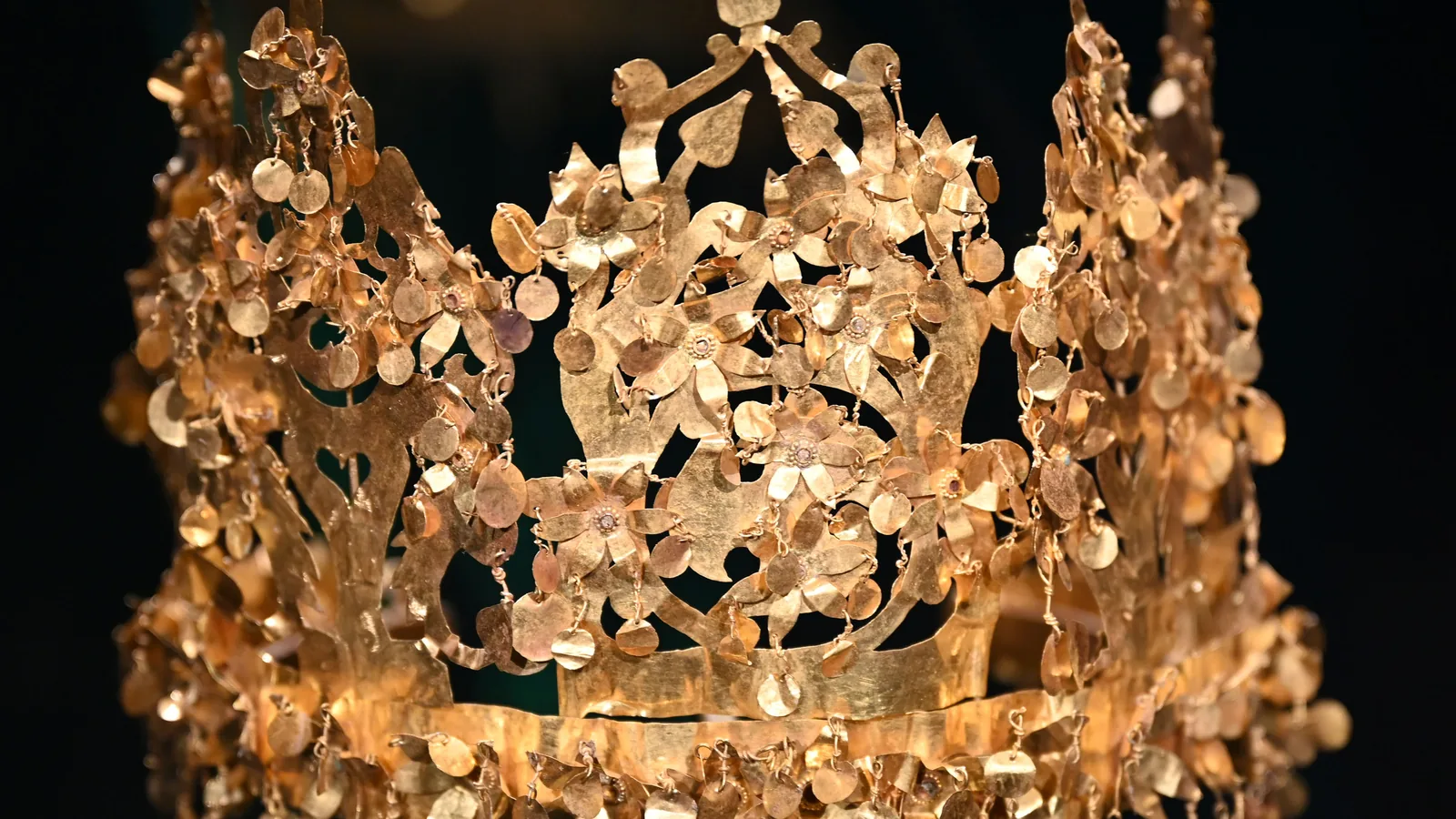 a gold crown of thin cut sheet made of five tree-like uprights hung with small gold discs and flowers, against a dark background