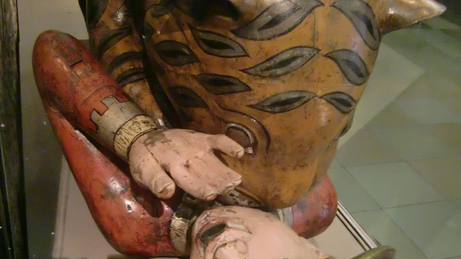 close view of a painted wooden tiger biting the neck of a carved man in a red coat, his hand raised to his face