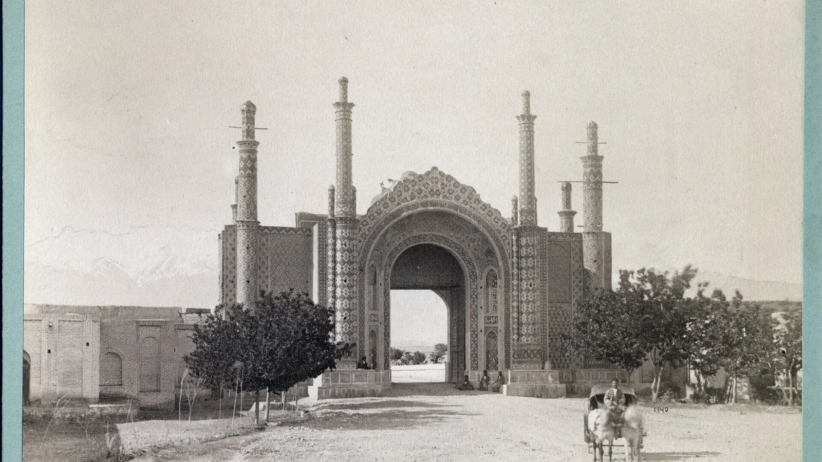 Sepia photograph of a tiled Qajar city gate with a central arch and flanking towers, an unpaved road in front