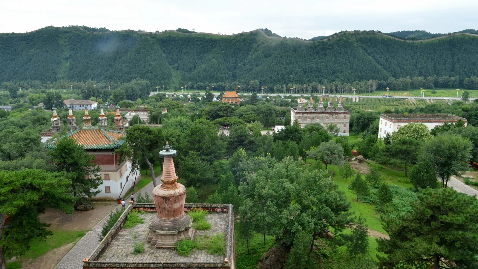 a view over wooded grounds to a red-walled temple complex with a white stupa in the foreground and green hills behind