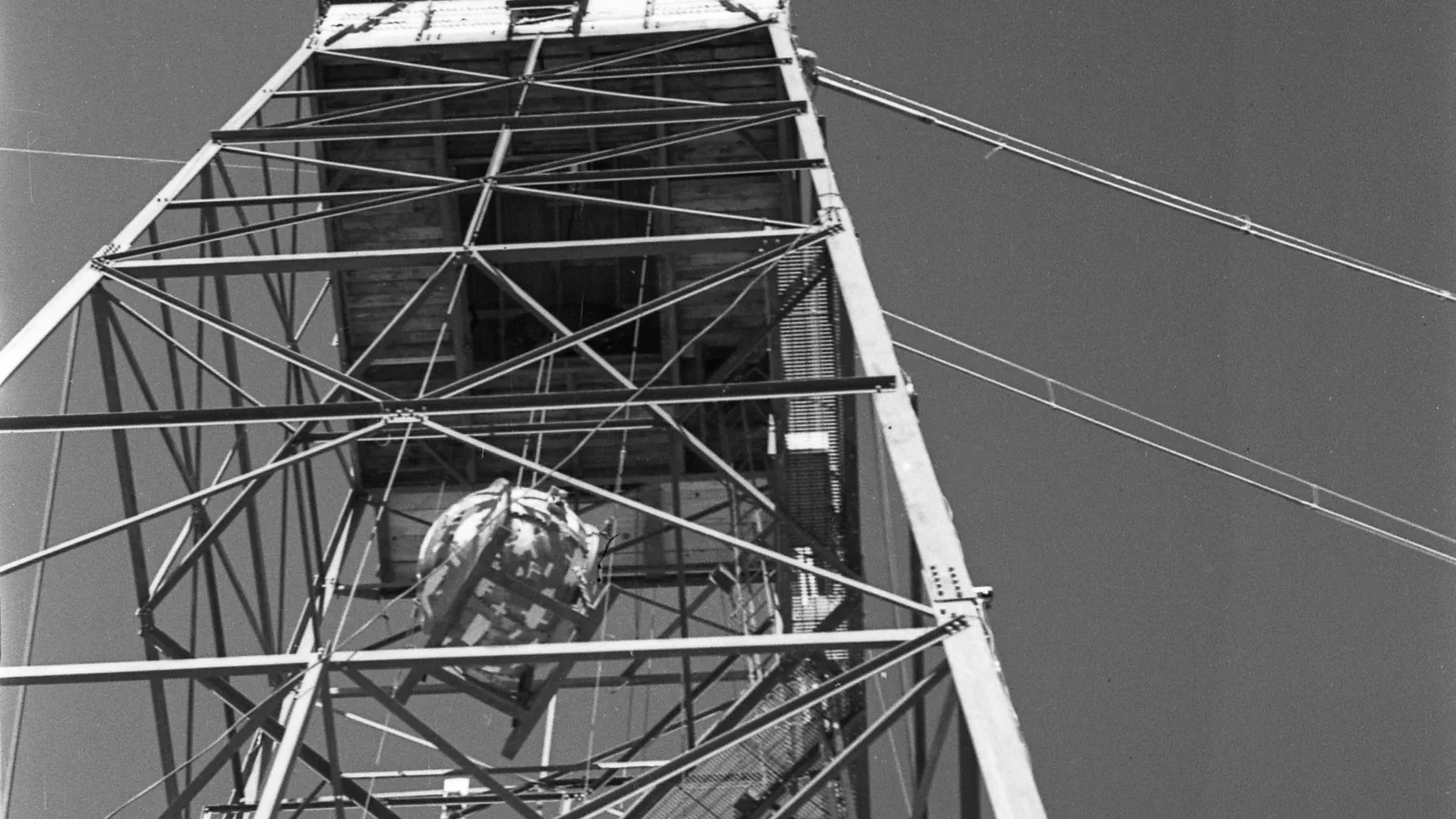 a black and white photograph looking up a steel lattice tower with a round device being hoisted inside it against the sky
