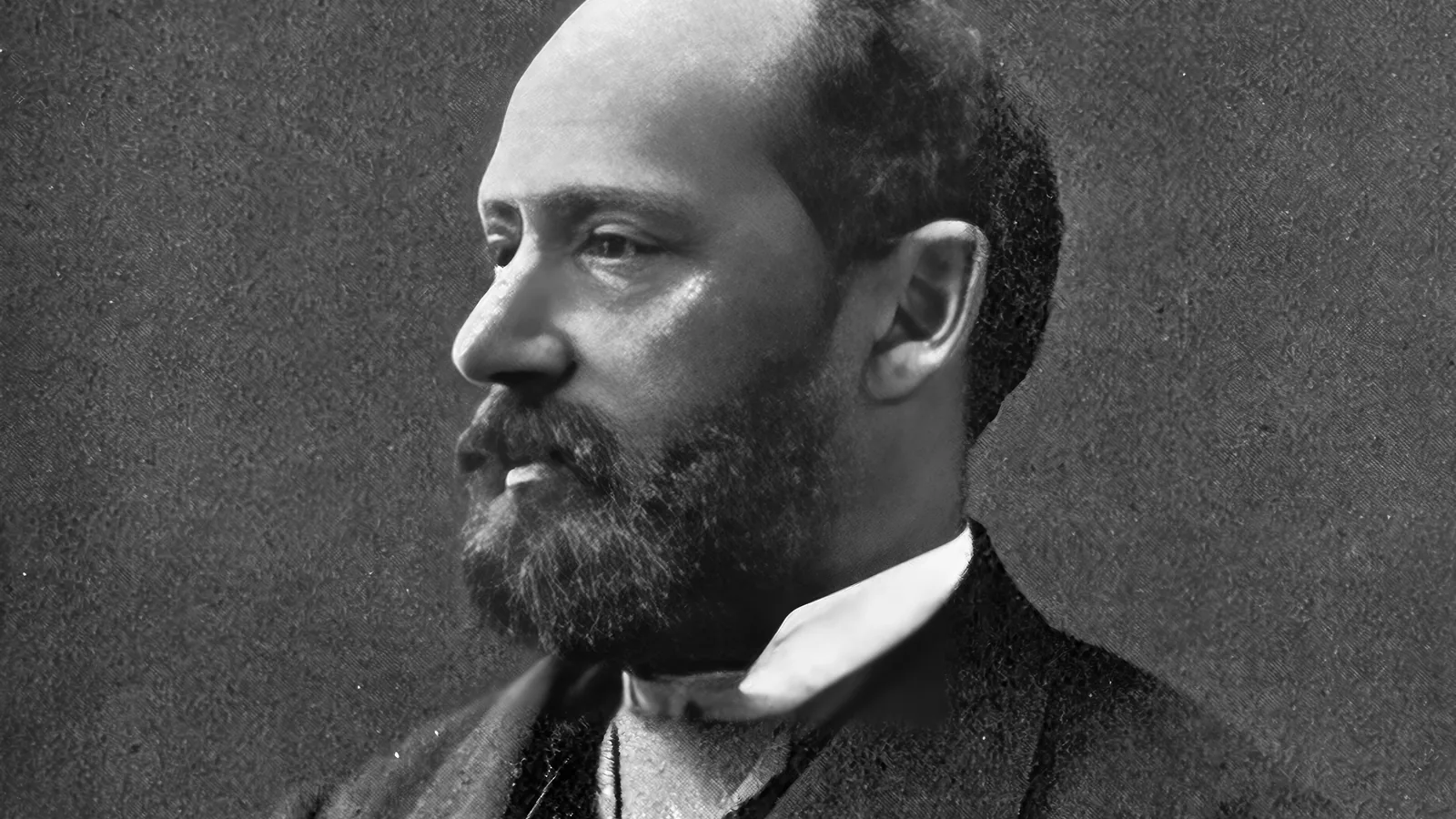 a black and white studio portrait of a bearded man in a dark coat and white collar, seated and looking to the side