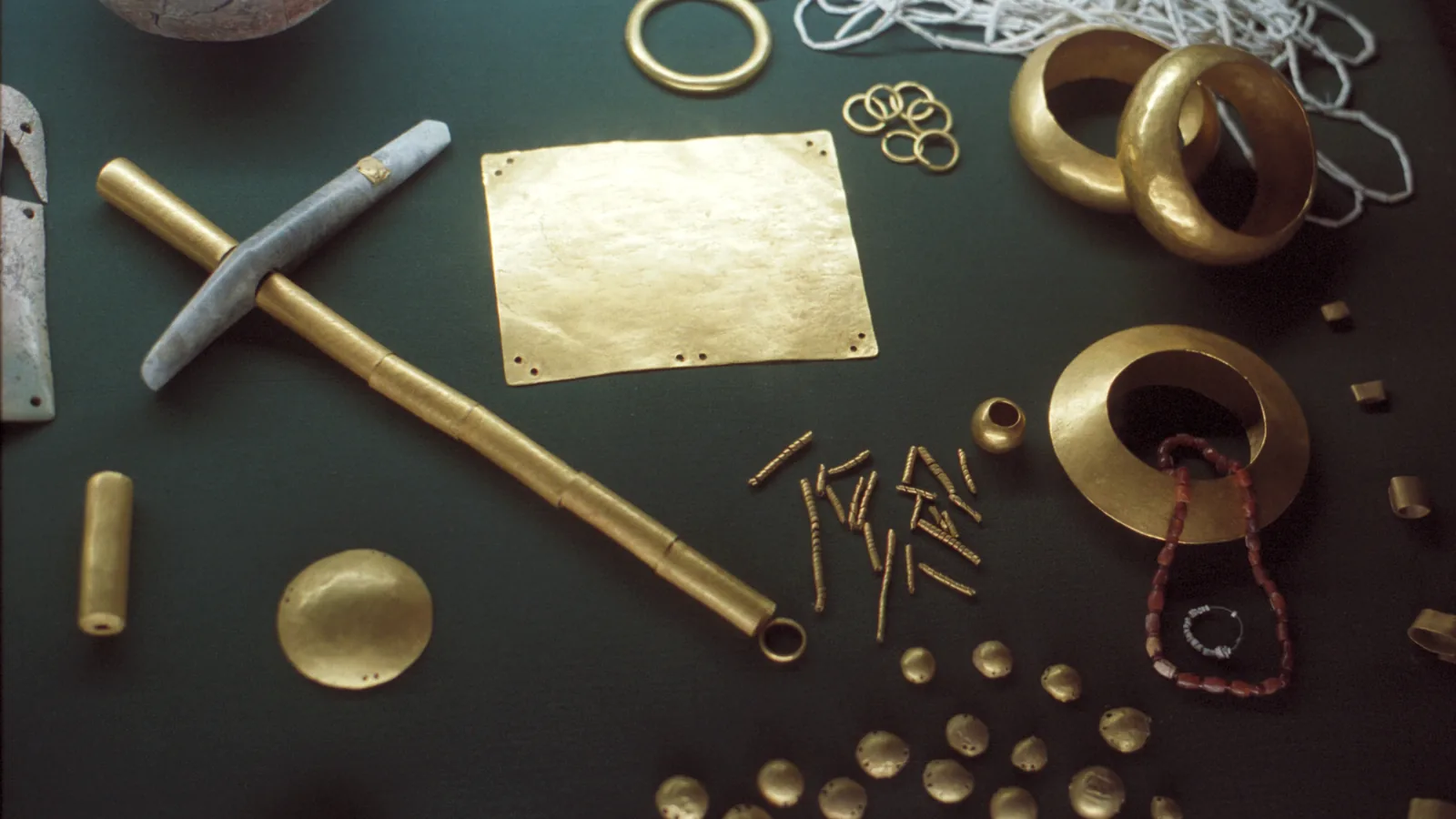Gold bracelets, rings, a stone-and-gold axe and small gold ornaments spread on a dark museum surface