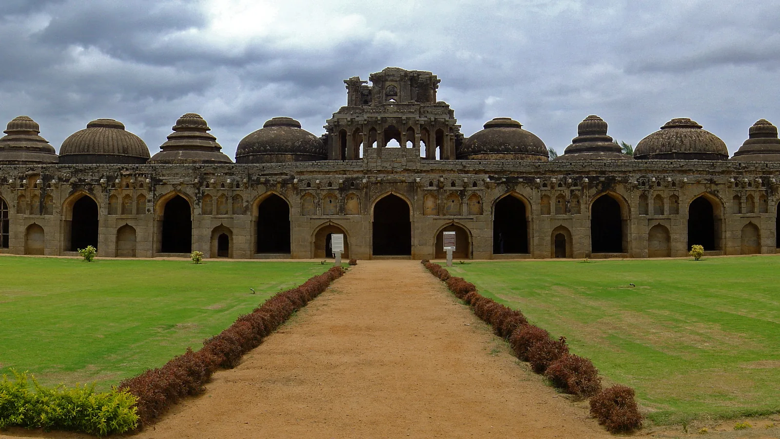 a long stone building with a row of arched doorways topped by domes of different shapes, on an open lawn
