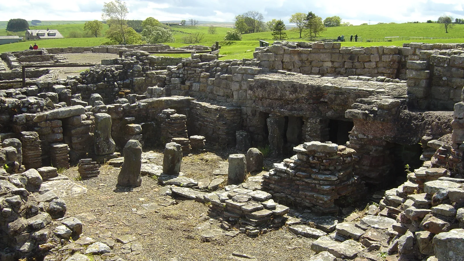 Excavated low stone walls of a Roman bathhouse in a grassy field, with rolling hills behind