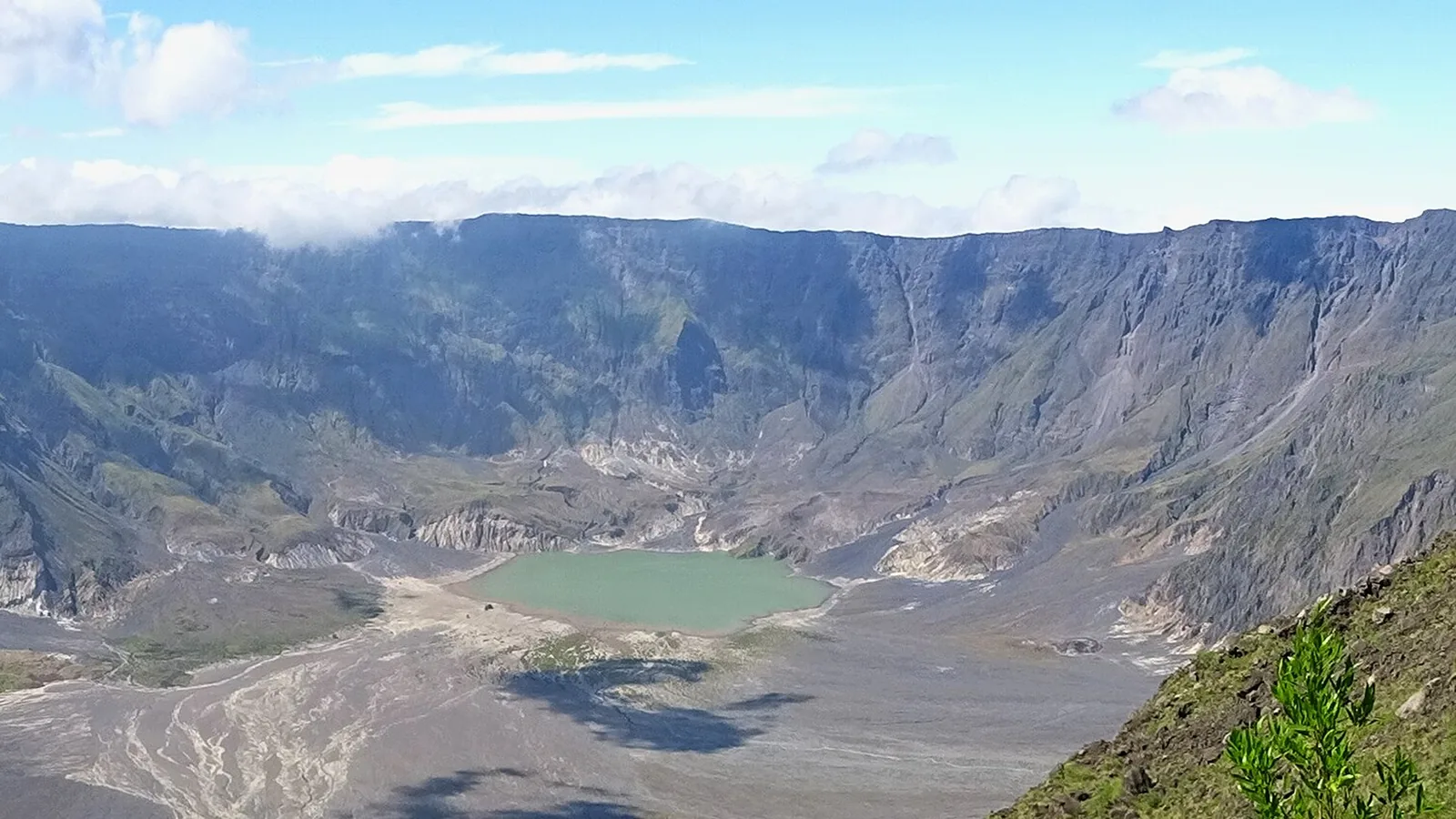 Wide ground-level panorama of the Tambora caldera floor with a turquoise crater lake and steep forested walls